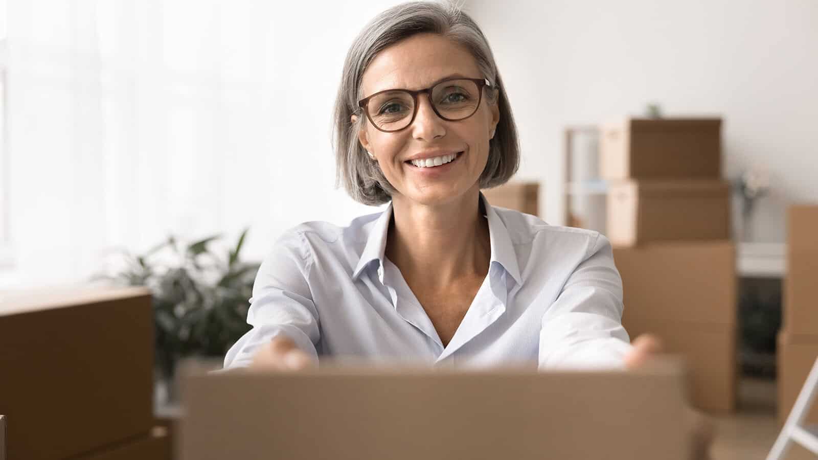 Smiling woman with gray hair and glasses holds a cardboard box, surrounded by more boxes in a bright room, suggesting unpacking or moving.