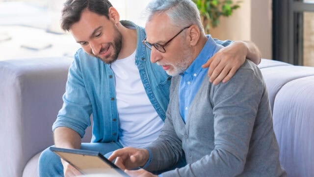Younger man and older man sit together on a couch, smiling as they look at a tablet. The younger man has his arm around the older man, who is wearing glasses and a gray sweater.