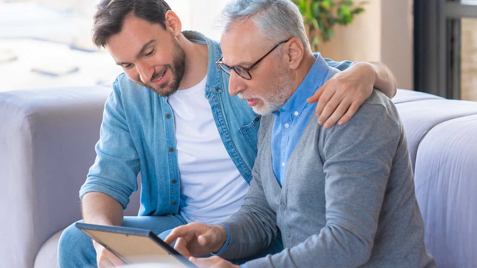 Younger man and older man sit together on a couch, smiling as they look at a tablet. The younger man has his arm around the older man, who is wearing glasses and a gray sweater.