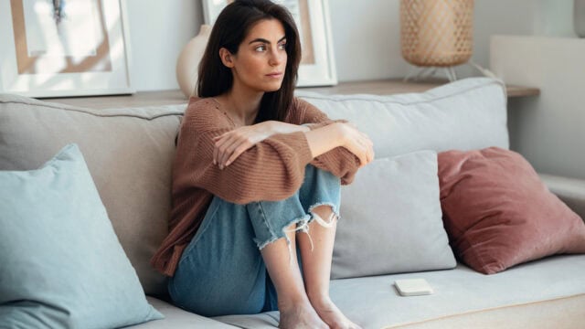 A woman sits on a couch with her knees pulled to her chest, looking thoughtful. She wears a brown sweater and ripped jeans. There are cushions and a phone beside her. The room is softly lit and decorated with framed art.