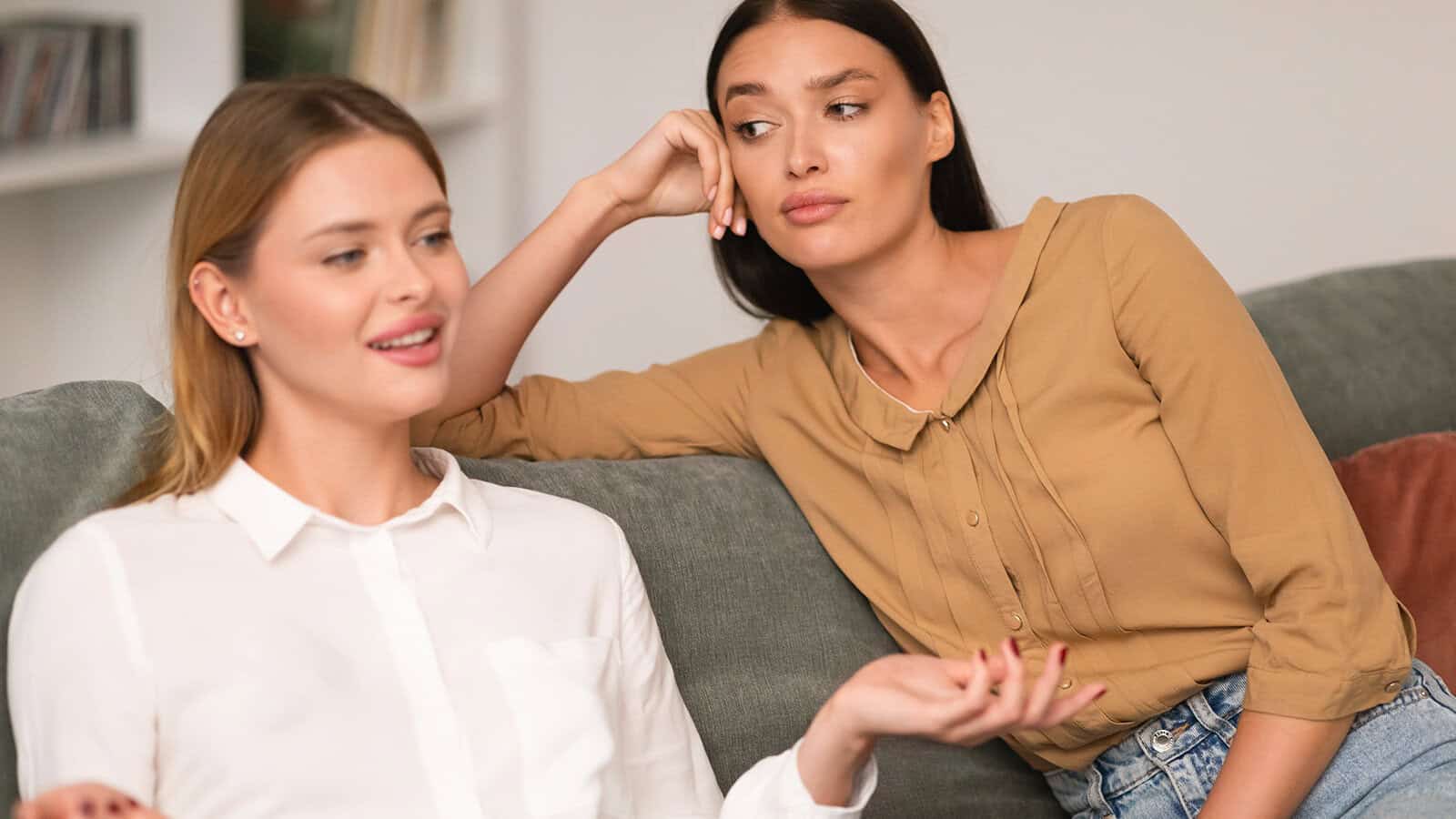 Two women sitting on a couch; one in a tan shirt listens with an unimpressed expression while the other, in a white blouse, talks animatedly with her hand raised.