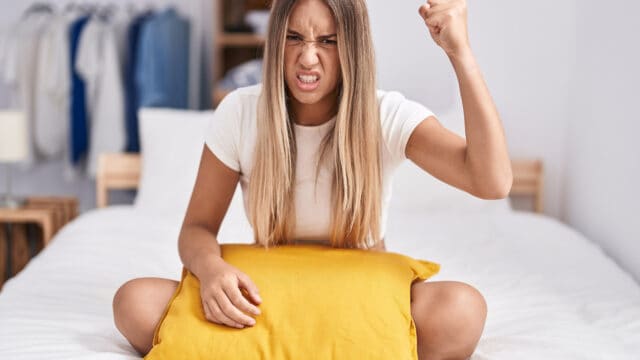 A young woman with long hair sits cross-legged on a bed, holding a yellow pillow and raising her fist with an angry facial expression. Clothing hangs in the background.