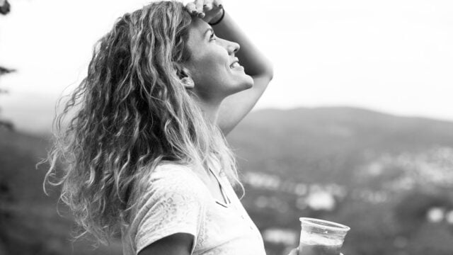 A woman with curly hair smiles and looks upward, shielding her eyes with one hand, while holding a plastic cup in the other. The background is an out-of-focus landscape. The image is in black and white.