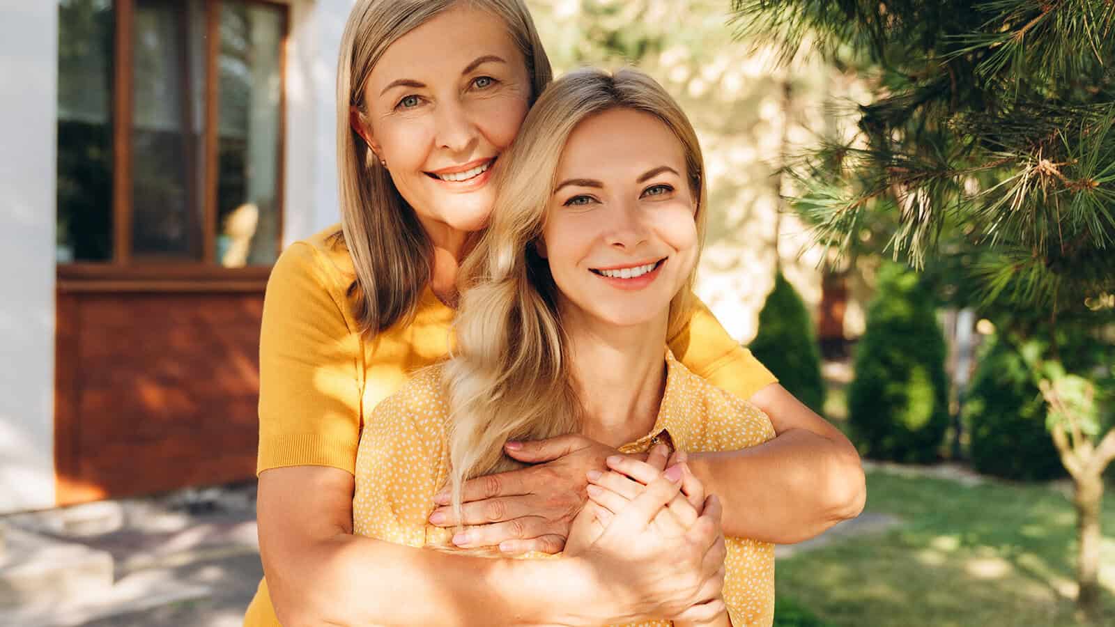 Two women, one older and one younger, smiling and hugging outdoors in a sunny garden. Both wear yellow tops and stand near a house and trees, radiating happiness and warmth.