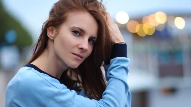 A woman with long brown hair wearing a light blue top stands outdoors, looking at the camera with a neutral expression. She is touching her hair, and there are blurred lights in the background.