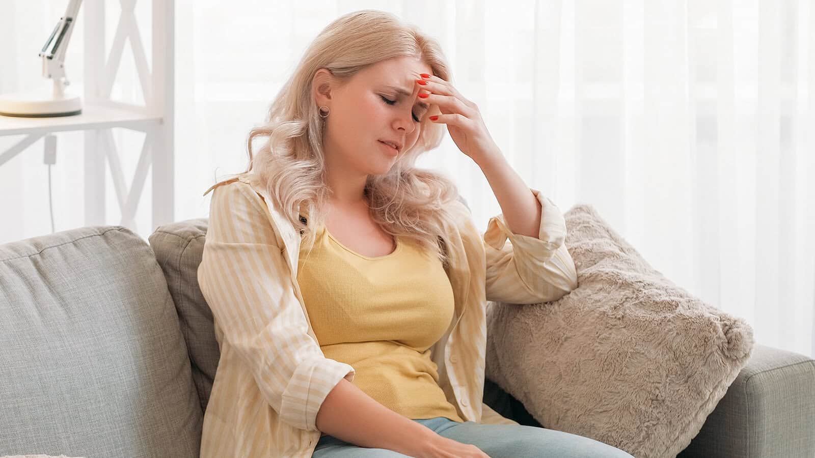 A woman with blonde hair sits on a sofa, wearing a yellow shirt and light striped jacket, holding her head in discomfort with her eyes closed, suggesting she might have a headache or feel unwell.