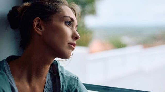 A woman with brown hair tied back gazes thoughtfully out of a window, soft daylight illuminating her face. She appears to be deep in thought, with a calm and introspective expression.