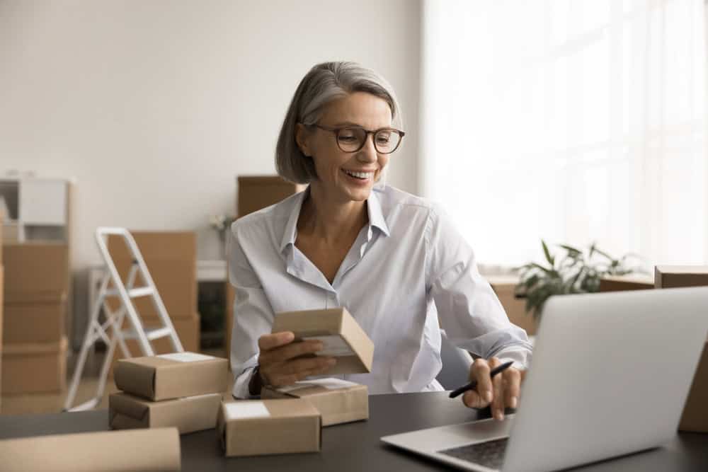 Smiling woman with gray hair and glasses holds a small package while working on a laptop at a desk surrounded by boxes, suggesting a home-based small business or online shop—perhaps offering proof of a life well-lived by giving things away and finding purpose through the psychology of sharing.