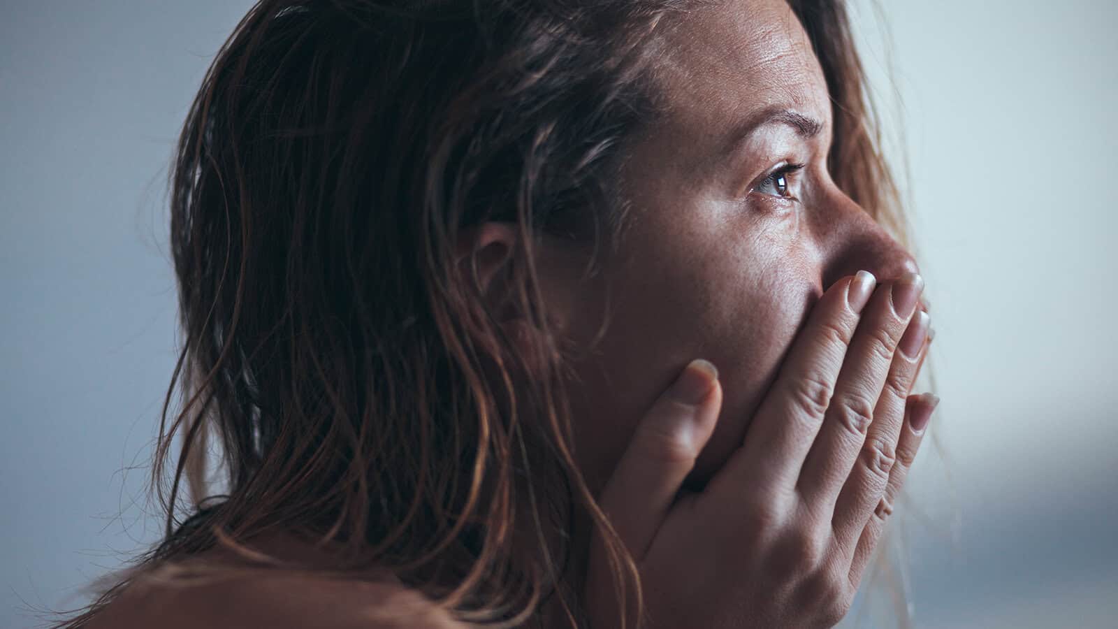 A person with wet hair covers their mouth with both hands, looking distressed or emotional. Their eyes are shiny, possibly with tears, and the background is blurred, focusing on the persons expression.