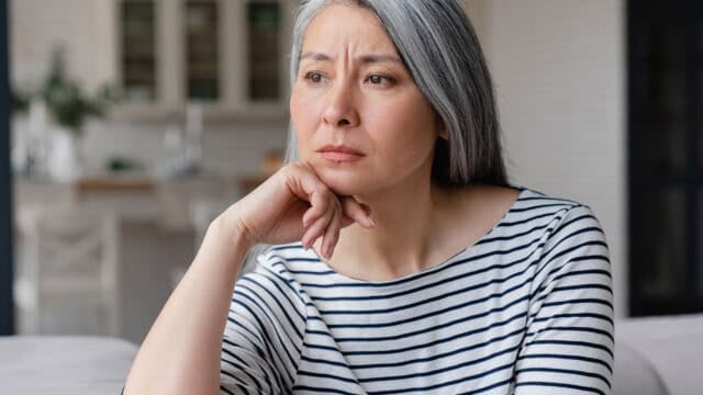 A woman with long gray hair and a striped shirt sits indoors, resting her chin on her hand, looking thoughtfully into the distance.