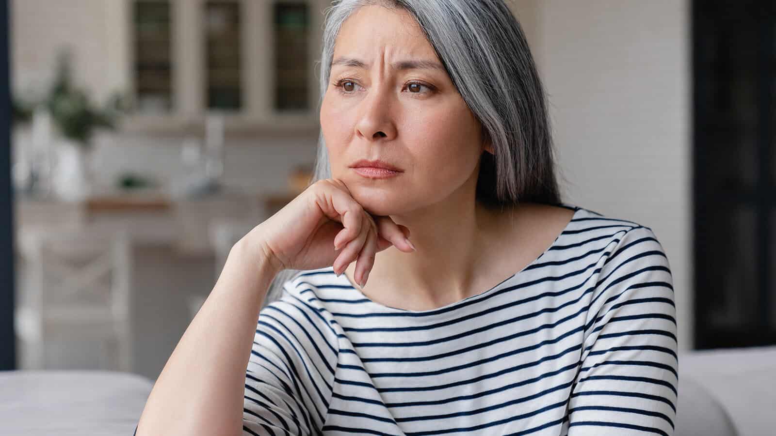 A woman with long gray hair and a striped shirt sits indoors, resting her chin on her hand, looking thoughtfully into the distance.