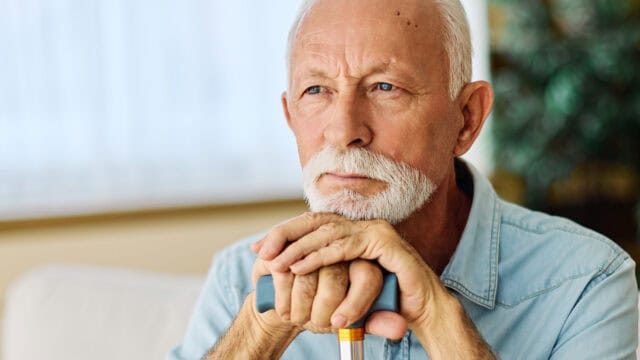 An elderly man with a white beard and mustache sits indoors, resting his hands on a cane and looking thoughtfully into the distance.