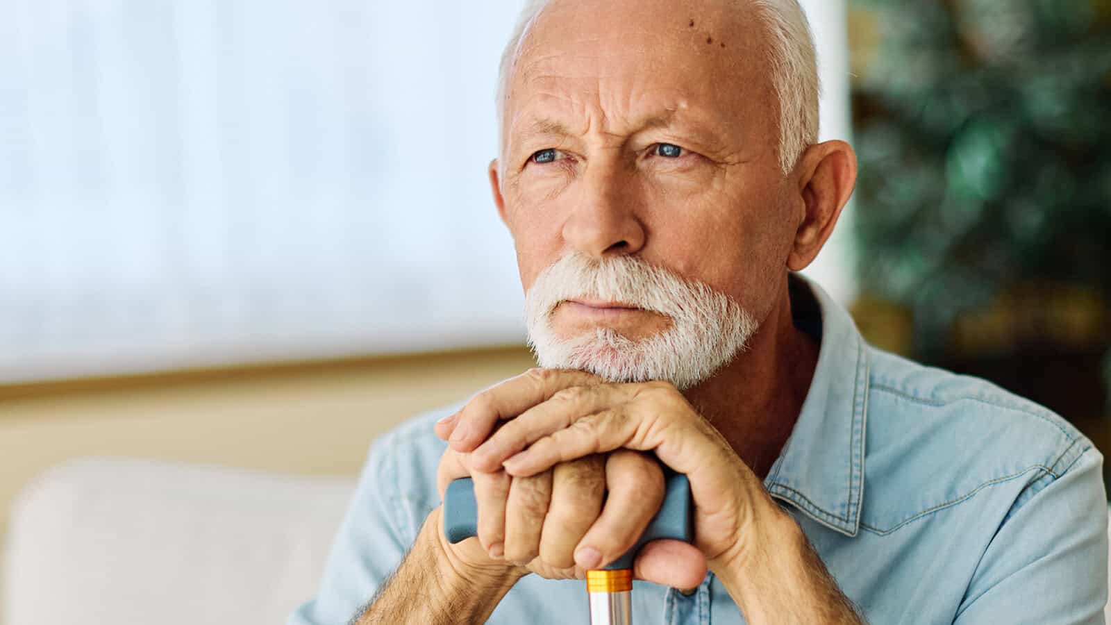 An elderly man with a white beard and mustache sits indoors, resting his hands on a cane and looking thoughtfully into the distance.