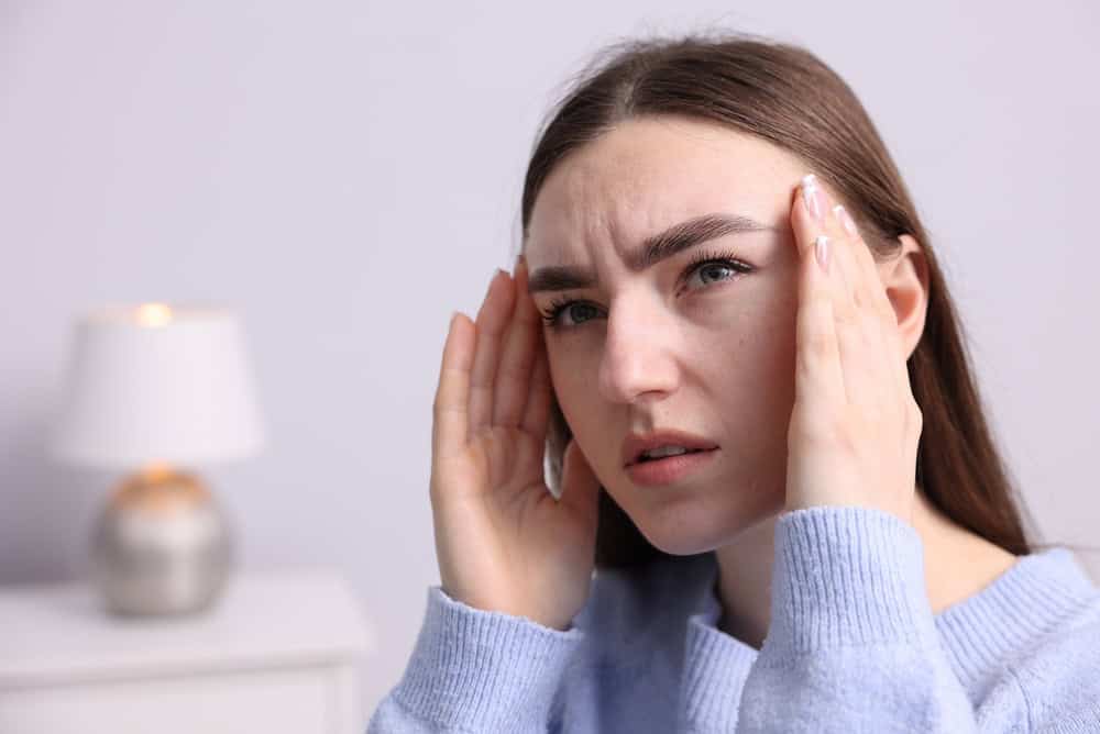 A woman with long brown hair holds her temples and frowns, appearing to have a headache; a blurred lamp is visible in the background, emphasizing the complex brain systems at play during moments of stress.