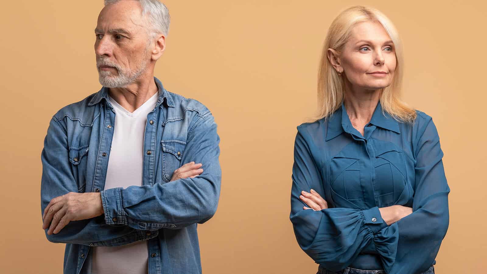 Older man and woman standing side by side with arms crossed, looking away from each other, both with serious expressions against a neutral beige background.