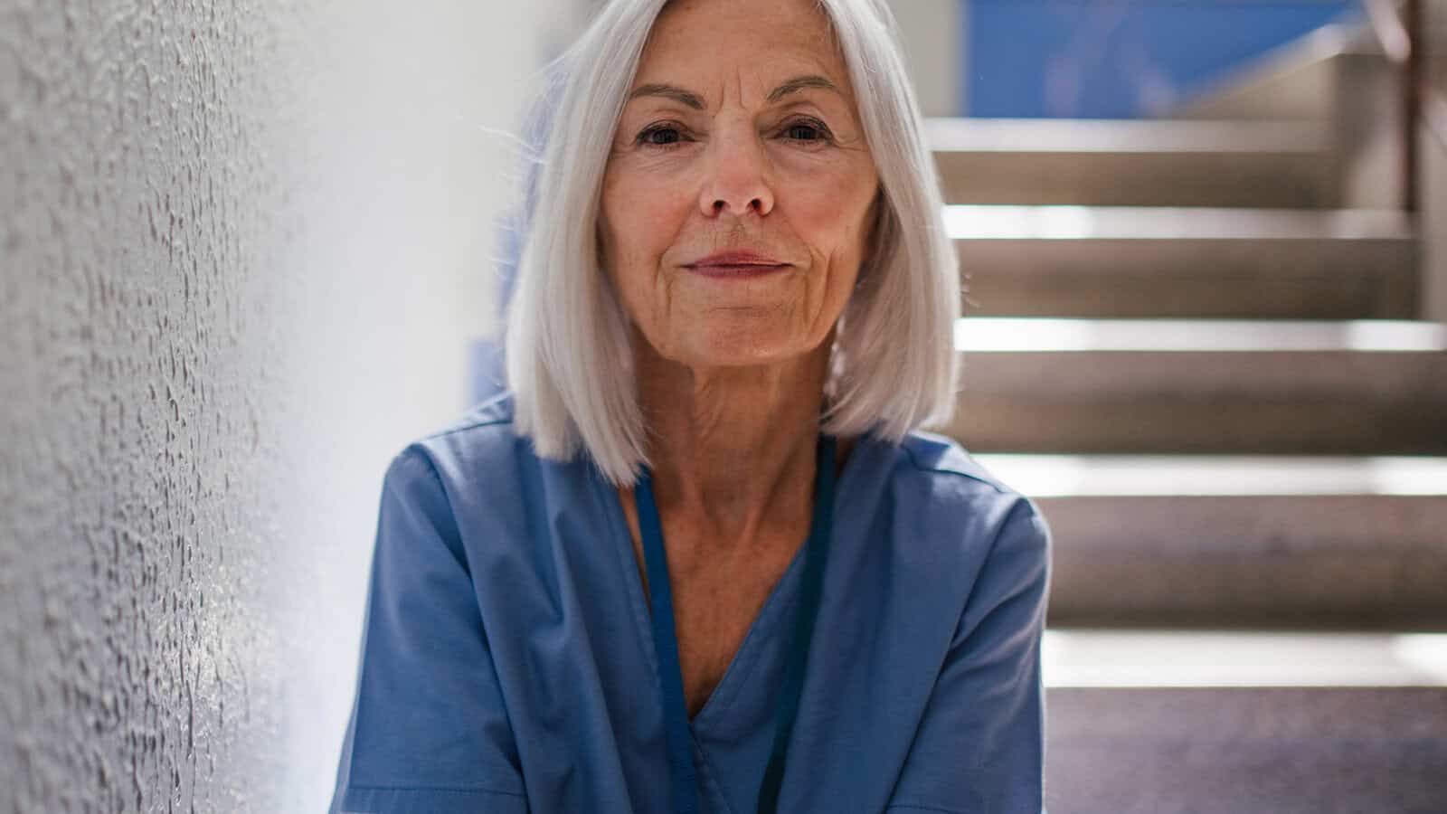 An older woman with shoulder-length gray hair wears blue scrubs and sits on stairs, looking confidently at the camera. Sunlight filters in from behind, casting soft light on her face.