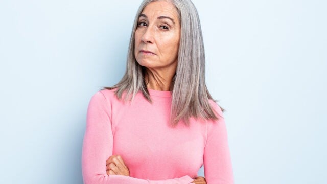 An older woman with long gray hair wearing a pink sweater stands against a light blue background, looking directly at the camera with a serious, slightly skeptical expression and her arms crossed.