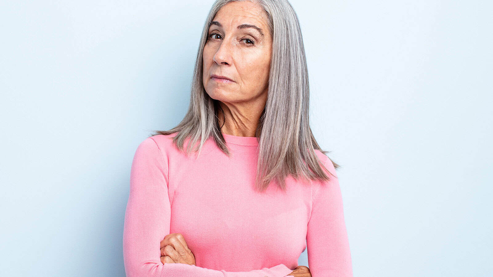 An older woman with long gray hair wearing a pink sweater stands against a light blue background, looking directly at the camera with a serious, slightly skeptical expression and her arms crossed.