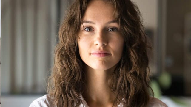 A young woman with long, wavy brown hair and a slight smile looks directly at the camera indoors, with a blurred background behind her.