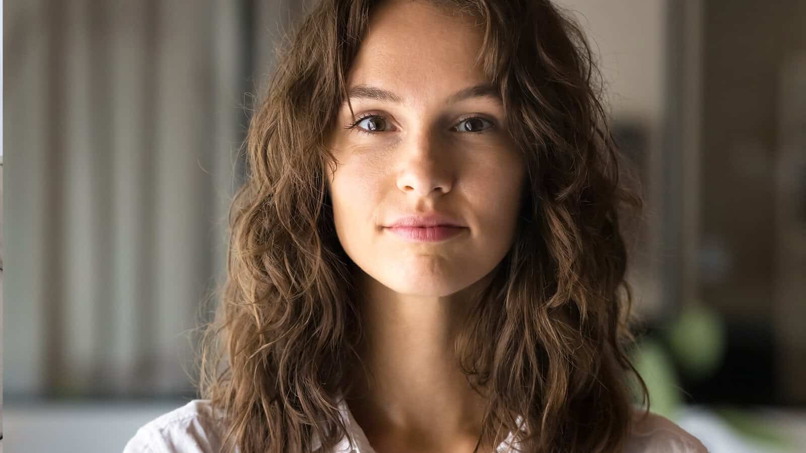 A young woman with long, wavy brown hair and a slight smile looks directly at the camera indoors, with a blurred background behind her.