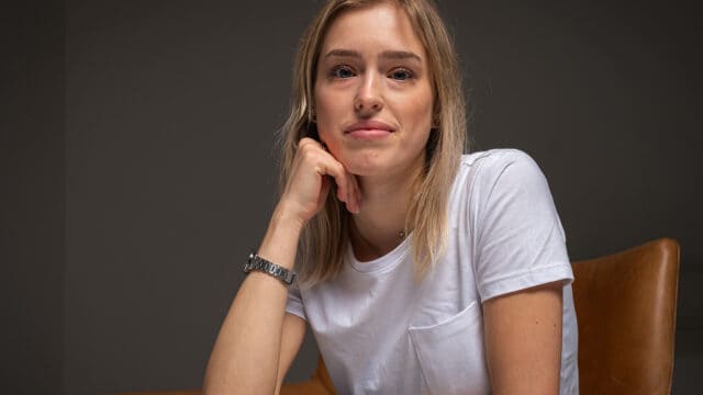 A woman with blonde hair sits on a brown chair, resting her chin on her hand. She wears a white t-shirt and a silver watch, looking calmly at the camera against a plain dark background.