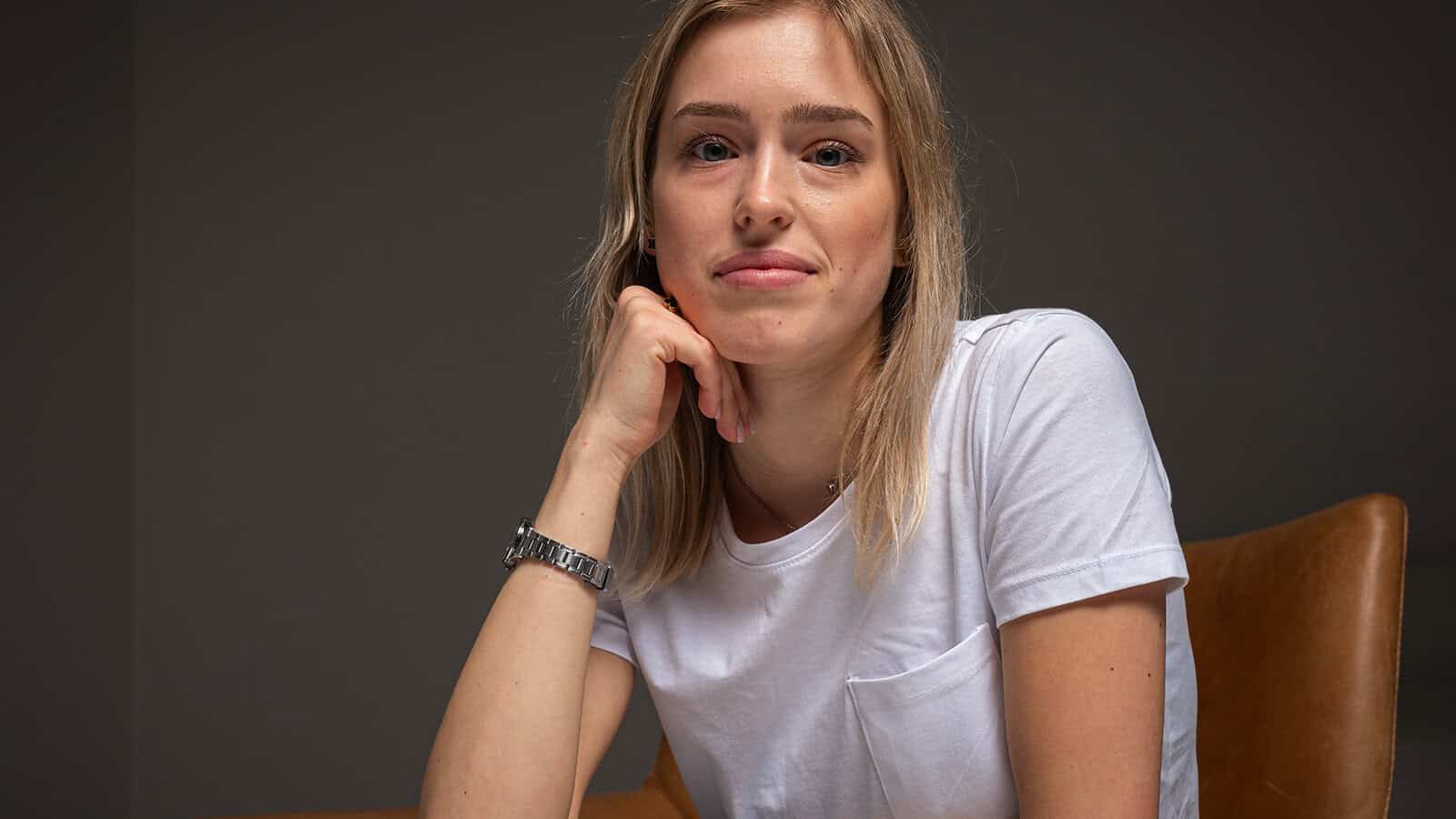 A woman with blonde hair sits on a brown chair, resting her chin on her hand. She wears a white t-shirt and a silver watch, looking calmly at the camera against a plain dark background.