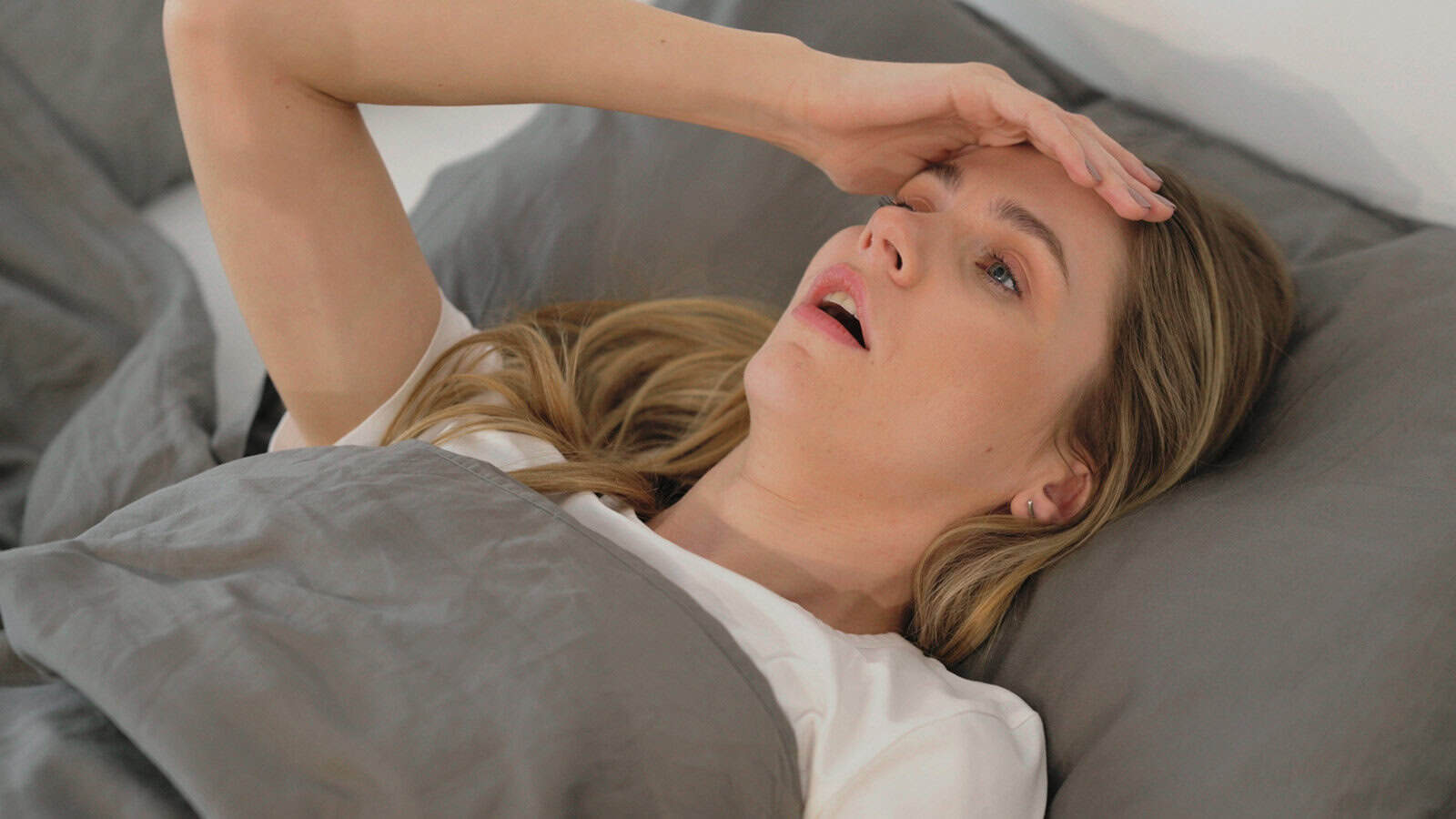 A woman lies in bed under gray sheets, looking distressed with her hand on her forehead and her mouth slightly open.