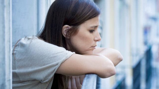 A woman with long brown hair leans on a railing, looking down with a thoughtful or sad expression. She wears a light-colored shirt and stands on an outdoor balcony or walkway.