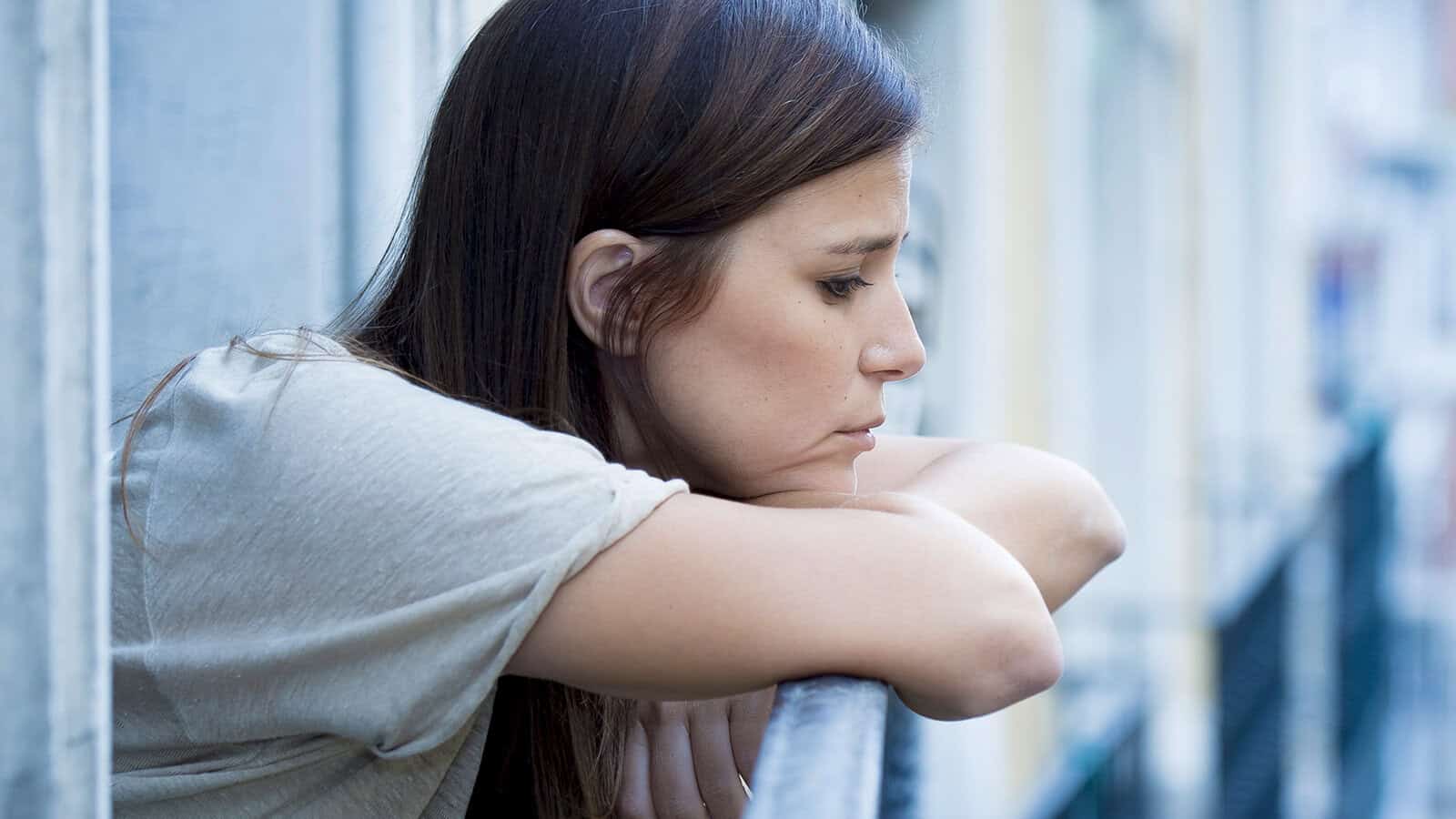 A woman with long brown hair leans on a railing, looking down with a thoughtful or sad expression. She wears a light-colored shirt and stands on an outdoor balcony or walkway.