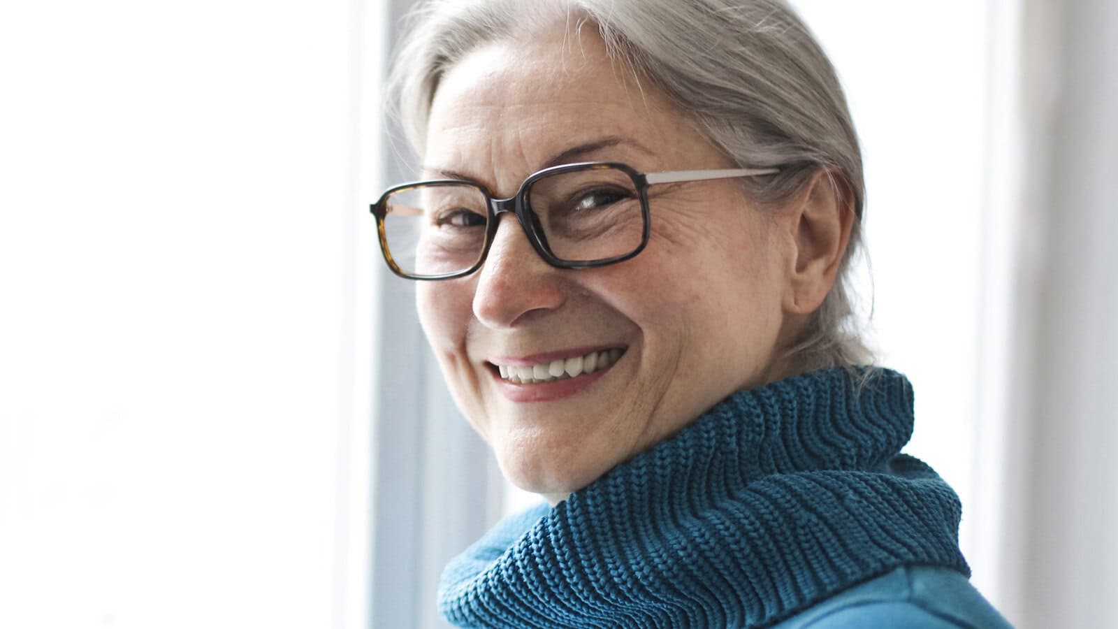 Smiling older woman with glasses and gray hair, wearing a blue turtleneck sweater, standing in front of a bright window.