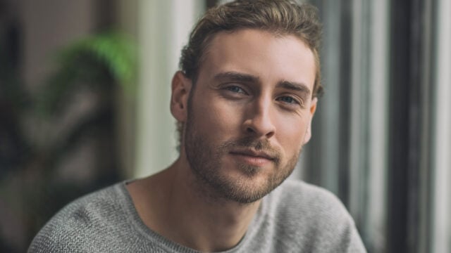 A man with light brown hair and a beard wears a gray sweater and looks into the camera, sitting indoors near a window with soft lighting and a blurred green plant in the background.