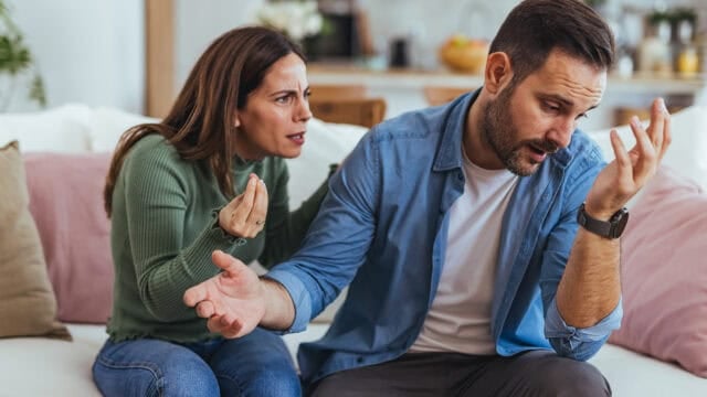 A woman appears frustrated while talking to a man who looks upset and is turning away on a couch in a home setting, highlighting the challenges of communication in relationships.