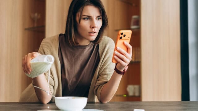 A woman pours milk into a bowl while looking at her phone with a surprised expression, sitting at a table in a modern kitchen, clearly distracted as her wandering mind shifts between breakfast and the unexpected news on her screen.