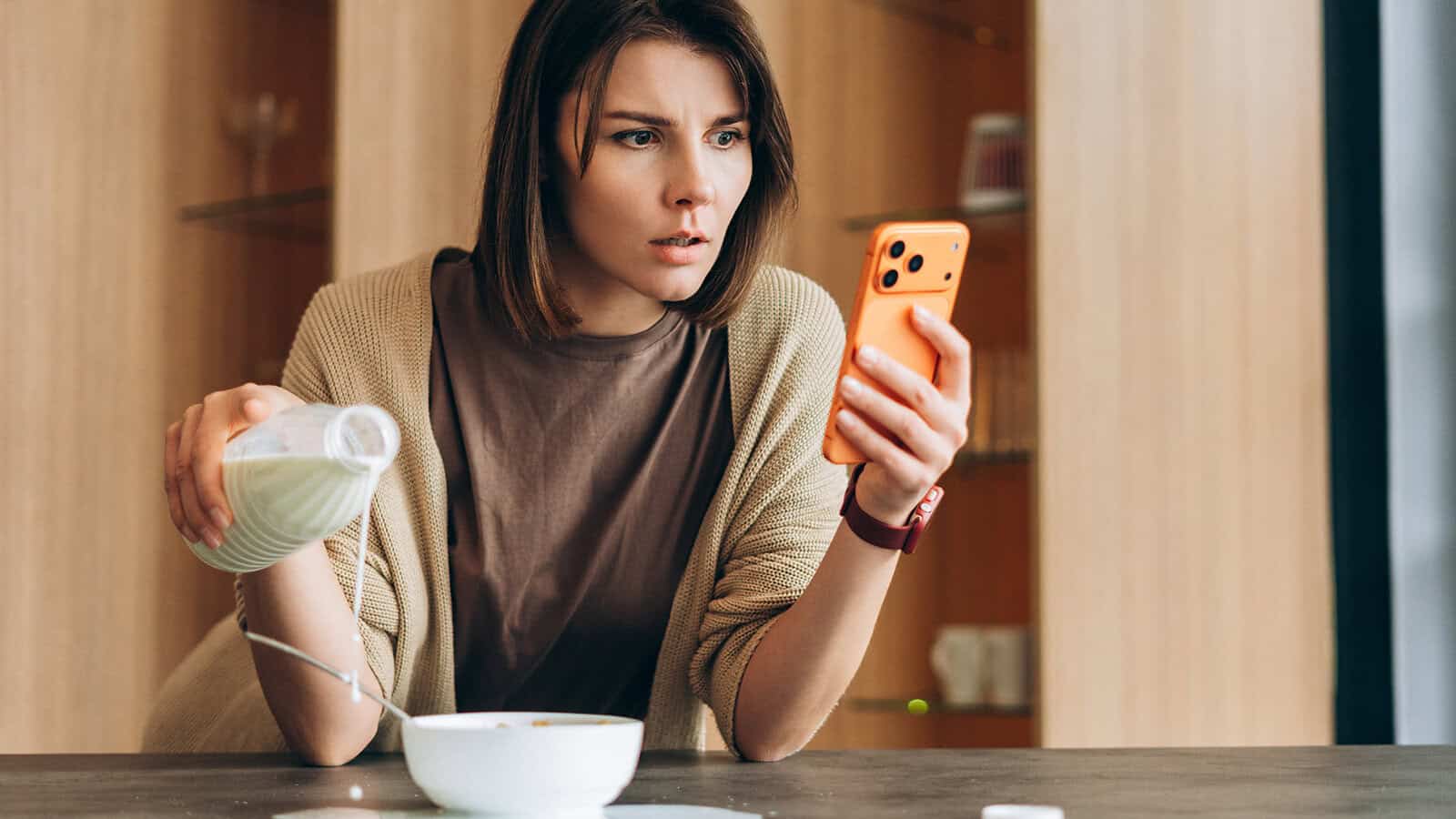 A woman pours milk into a bowl while looking at her phone with a surprised expression, sitting at a table in a modern kitchen, clearly distracted as her wandering mind shifts between breakfast and the unexpected news on her screen.