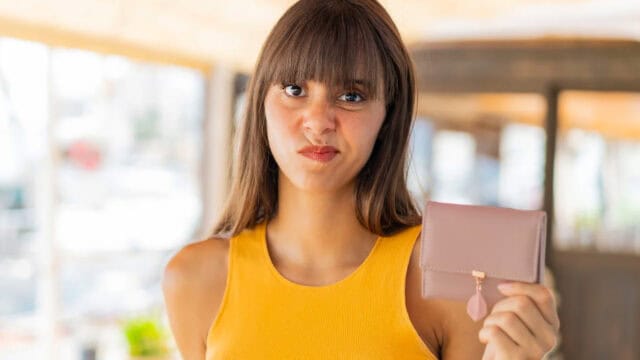 A woman in a yellow sleeveless top holds up a closed pink wallet and makes a displeased facial expression, standing indoors with a blurred background—capturing the anxiety that can come from Gen Z money habits and modern financial habits.