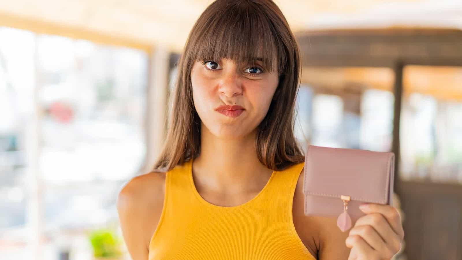 A woman in a yellow sleeveless top holds up a closed pink wallet and makes a displeased facial expression, standing indoors with a blurred background—capturing the anxiety that can come from Gen Z money habits and modern financial habits.