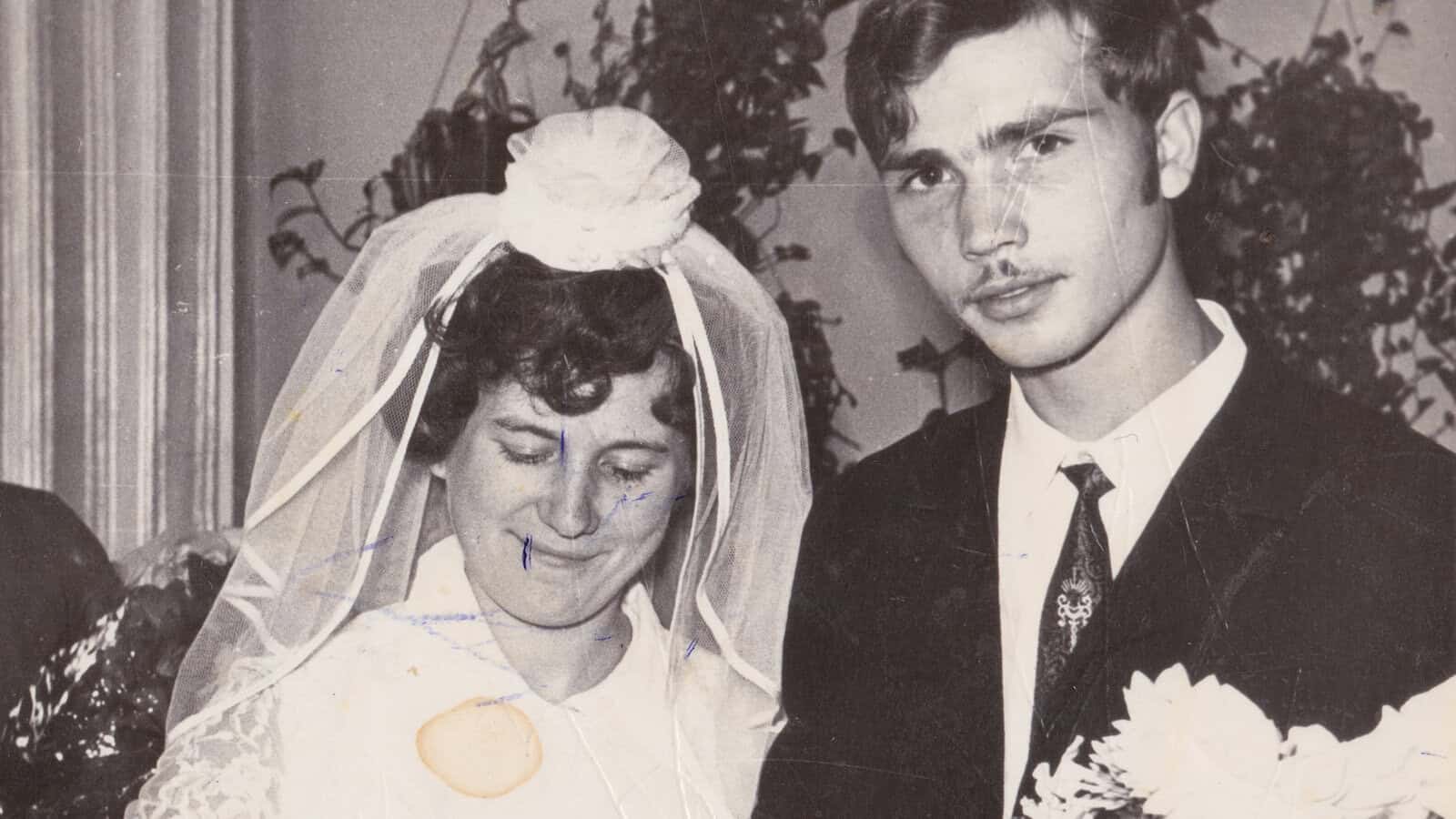 A vintage black-and-white photo of a bride in a veil and a groom in a suit, both standing close together and holding flowers, with visible age marks and creases on the image—a timeless snapshot capturing the essence of a good marriage that still inspires Millennials and Gen Z today.