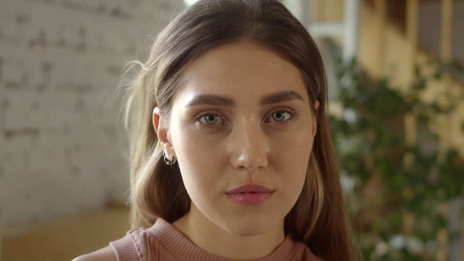 A young woman with long light brown hair and blue eyes, representing the style of many Millennials, looks directly at the camera with a neutral expression, standing indoors with soft lighting and a blurred background.