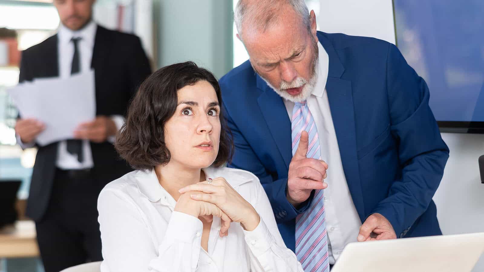 A woman at a desk looks anxious as an older man in a suit gestures and speaks to her. Another man in the background—perhaps a good boss admired by Gen Z and Millennials—holds papers, watching the interaction.