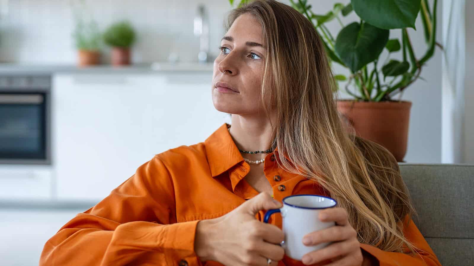 A woman in an orange shirt sits indoors, holding a mug and looking thoughtfully into the distance, perhaps reflecting on how best to care for her mother. There are green potted plants and a kitchen visible in the blurred background.