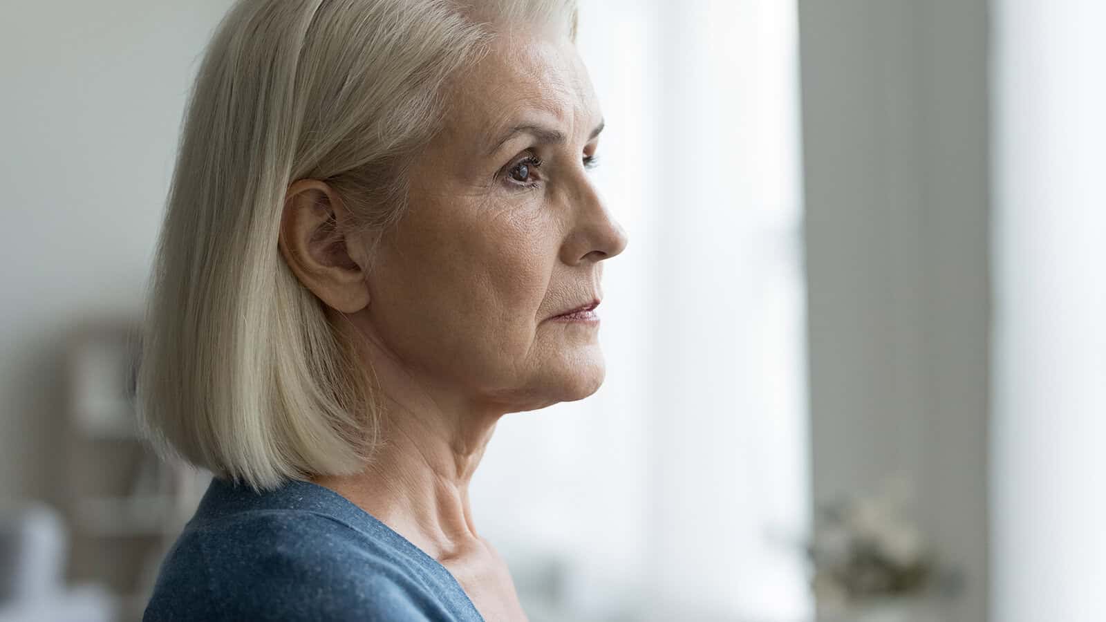 Elderly woman with short gray hair and a blue sweater gazes thoughtfully out of a bright window, the soft natural light illuminating her face as she reflects on unmarked milestones and cherished family memories.