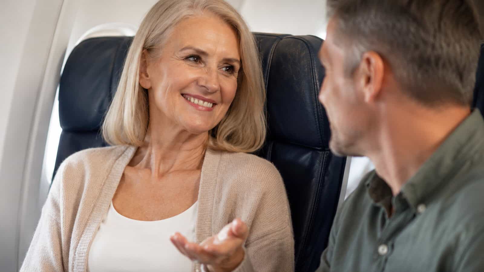 Smiling older woman, embodying the friendly traits often seen in older people, talks to a man while sitting together on an airplane; both appear relaxed and engaged as they chat with strangers during their journey.