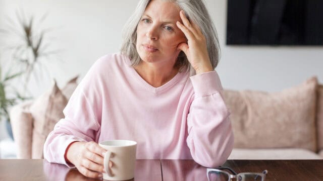 A woman with gray hair, wearing a pink sweater, sits at a table holding a white mug. She looks thoughtful or concerned, resting her head on one hand—perhaps contemplating parenting tips or signs to watch for as her adult child is struggling. A pair of glasses and a couch are in the background.