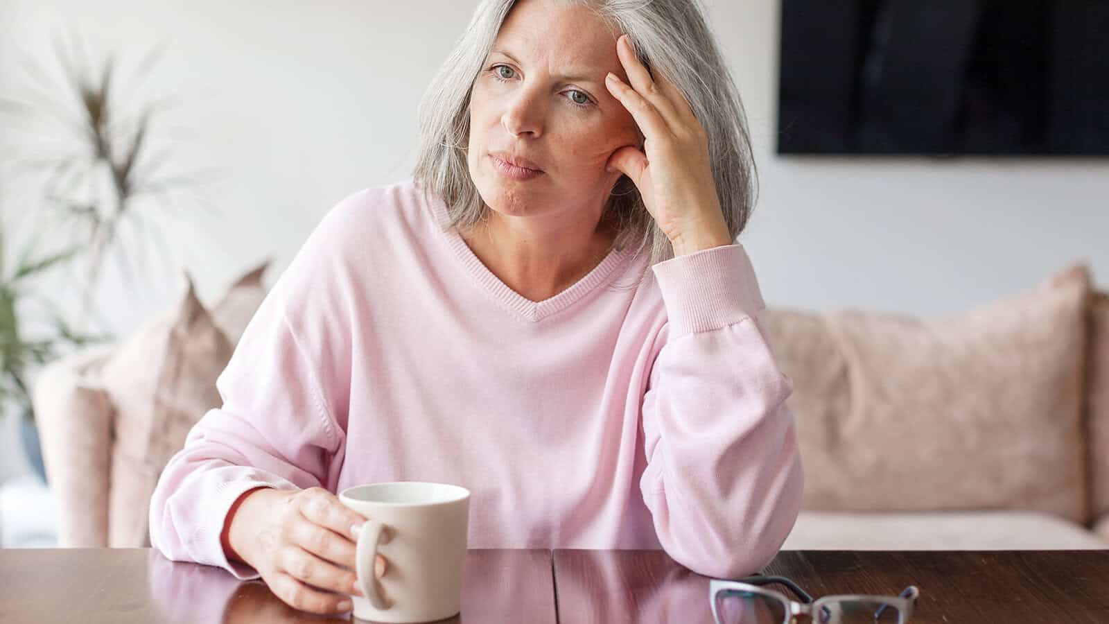 A woman with gray hair, wearing a pink sweater, sits at a table holding a white mug. She looks thoughtful or concerned, resting her head on one hand—perhaps contemplating parenting tips or signs to watch for as her adult child is struggling. A pair of glasses and a couch are in the background.