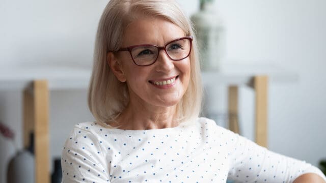 Smiling older woman with straight, shoulder-length gray hair and glasses, wearing a white shirt with blue polka dots, sitting indoors with a blurred background—a perfect example of people in their 70s who have dropped habits that no longer serve them and stopped explaining themselves to others.