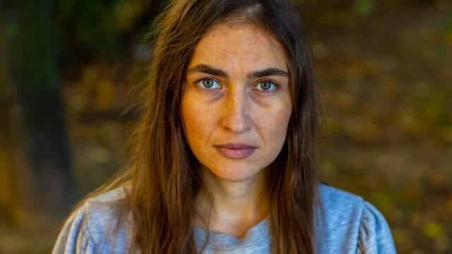A woman with long brown hair and green eyes gazes seriously at the camera, wearing a gray shirt. The background is blurred, showing outdoor foliage with earthy tones. Her intense expression suggests she’s just heard one of those infuriating phrases that can shut down conversation or trigger a victim mentality.