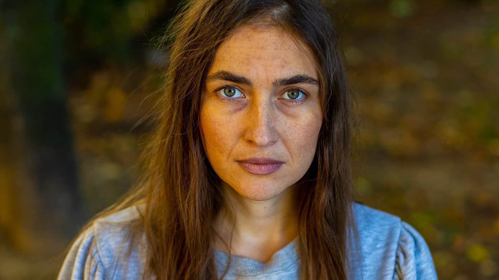 A woman with long brown hair and green eyes gazes seriously at the camera, wearing a gray shirt. The background is blurred, showing outdoor foliage with earthy tones. Her intense expression suggests she’s just heard one of those infuriating phrases that can shut down conversation or trigger a victim mentality.