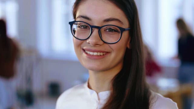 A young woman with long brown hair and glasses smiles warmly at the camera indoors, her easygoing demeanor evident against a softly blurred background suggesting a bright, modern setting.