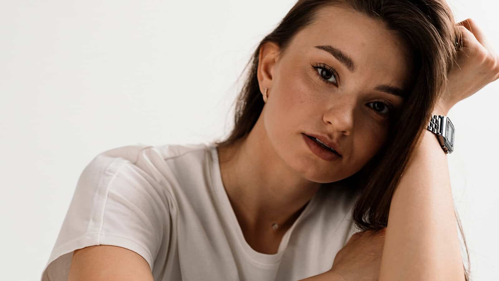 A woman with long brown hair and a white t-shirt rests her head on her arm, looking into the camera with a neutral expression against a plain light background. Her calm gaze suggests someone who remains emotionally steady, even when others might fall apart in crisis.