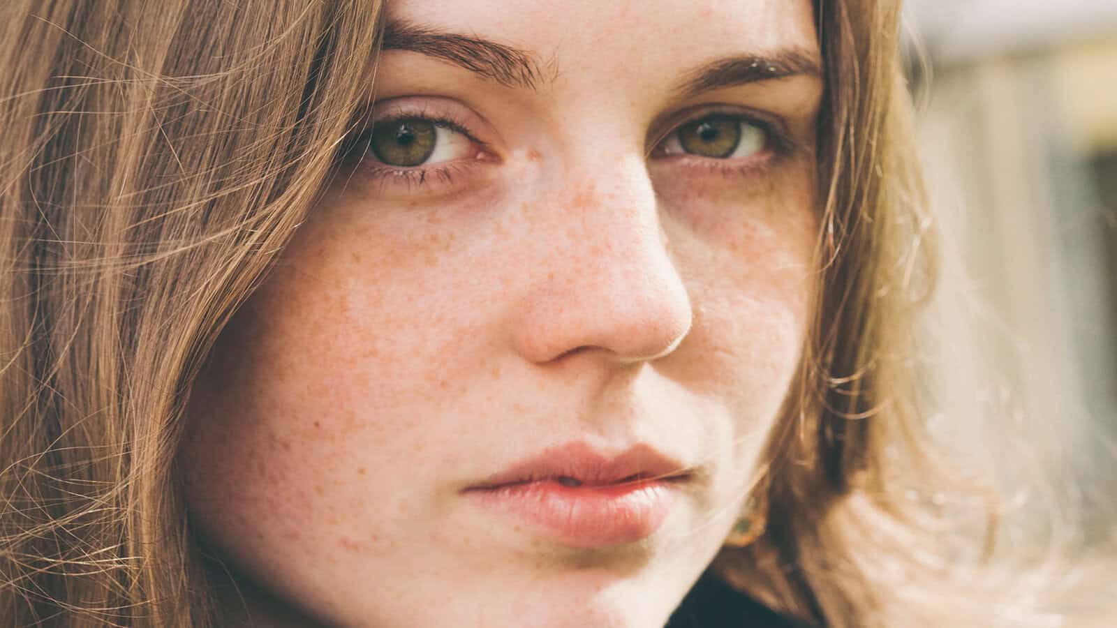 A close-up of a young person with light skin, green eyes, freckles, and brown hair, looking directly at the camera with a neutral expression—an air that hints at stubborn habits and a quiet determination to refuse to change.
